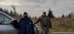 Policjantka umundurowana wraz ze strażnikiem leśnym w mundurze stoją przy samochodzie terenowym. W tle widać drzewa i krzaki. Patrol ma maseczki na twarzy. Na zdjęciu jest pora dzienna Na dole zdjęcia w prawym rogu jest umieszczony napis www.dolnoslaska.policja.gov.pl.