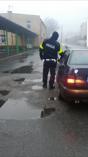 Policjant umundurowany stoi na drodze przy samochodzie osobowym i przez okno podje alkotest kierującemu. Po lewej stronie widać wiatę przystanku autobusowego i zabudowania