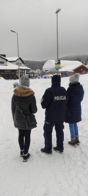 Policjant w mundurze oraz dwie kobiety idą po zaśnieżonej drodze dookoła leży śnieg Na zdjęciu widać  zabudiowania.