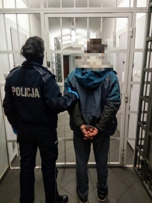 Na zdjęciu umundurowany policjant prowadzi zatrzymanego mężczyznę, który ma założone kajdanki na rękach do tyłu. Zdjęcie wykonane w pomieszczeniu , widać w tle kraty.