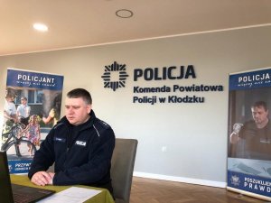 Policjant umundurowany siedzi przy laptopie, który znajduje sie na stole .Z tyłu znajduje sie napis Komenda Powiatowa Policji w Kłodzku. Obok stoi baner dotyczący rekrutacji do Policji.. Zdjęcie wykonane w pomieszczeniu sali sztabowej.