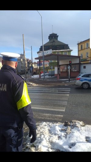 Umundurowany policjant Ruchu Drogowego stoi przy przejściu dla pieszych. Policjant stoi tyłem , w tle widać zabudowania oraz leżący na chodniku śnieg. Zdjęcie zrobione w porze dziennej.