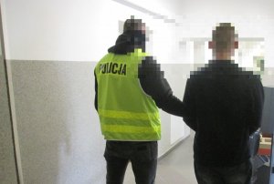 Policjant operacyjny w kamizelce z napisem Policja prowadzi zatrzymanego mężczyznę. W tle widać korytarz. Zdjęcie zrobione w pomieszczeniu służbowym.