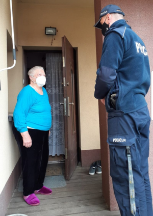 Policjant w mundurze i maseczce na twarzy stoi przy otwartych drzwiach do domu obok niego stoi starsza kobieta ubrana w sweter, spodnie oraz kapcie. Kobieta  na twarzy ma maseczkę.