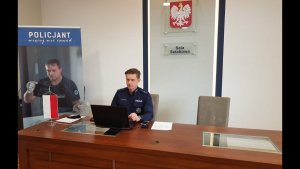 Policjant w mundurze siedzi przy stoliku na którym znajduje się laptop. Za policjantem na ścianie znajduje sie napis Sala Sztabowa oraz godło państwowe.PO lewej stronie widać baner dot.rekrutacji do Policji z Napisem Policjant więcej niż zawód z wizerunkiem policjanta na nim.