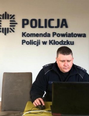 Umundurowany policjant siedzi przy stoliku na którym znajduje  się otwarty laptop.Widać na ścianie napis Policja Komenda Powiatowa Policji w Kłodzku.Zdjęcie wykonane w pomieszczeniu