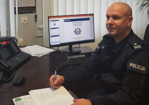 Komendant Powiatowy Policji w Kłodzku podinspektor Rafał Siczek siedzi przy biurku, a z prawej jego strony znajduje się laptop na ekranie którego widać slajd o treści odprawa roczna komendy Powiatowej Policji w Kłodzku