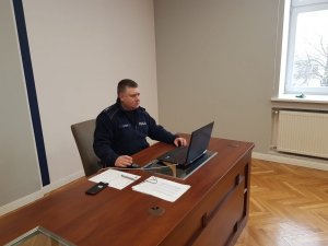 Umundurowany policjant siedzi przed stolikiem, na którym jest laptop oraz dokumenty  .Zdjecie wykonane w porze dziennej w pomieszczeniu służbowym -sala sztabowa kłodzkiej policji