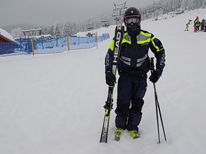 Policjant stoi w pobliżu stoku, trzyma w ręku narty , w tle widać stok oraz narciarzy