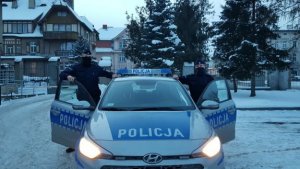 dwóch umundurowanych policjantów stoi po lewej i prawej stronie radiowozu policyjnego oznakowanego. W tle widać śnieg i ogrodzenie.Zdjęcie zrobione na świeżym powietrzu. W radiowozie włączone są światłą.