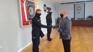 dwóch policjantów stoi w sali sztabowej i przyjmują od komendanta akt mianowania  w tle widać w gablocie sztandar policji powiatu kłodzkiego