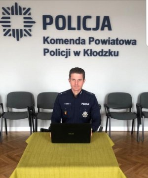 Policjant w mundurze siedzi przed stolikiem na którym znajduje sie laptop. W tle widać napis Komenda Powiatowa Policji w Kłodzku. Zdajecie zrobione w sali sztabowej kłodzkiej policji