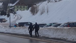 dwóch policjantów idzie chodnikiem, dookoła lezy śnieg.Po prawej ich stronie są zaparkowane samochody