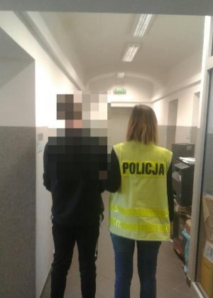 Policjantka prowadzi zatrzymanego w pomieszczeniach służbowych-korytarz. policjantka ma na sobie kamizelkę z napisem policja