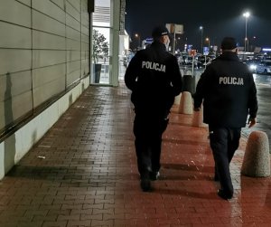 Dwóch umundurowanych policjantów idzie w kierunku galerii handlowej. W tle widać zaparkowane samochody na parkingu