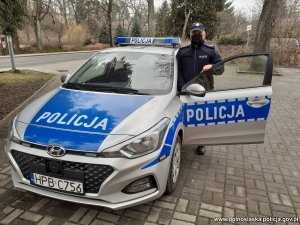 Policjant z maseczką na twarzy w mundurze stoi przy radiowozie oznakowanym w tle widać drzewa