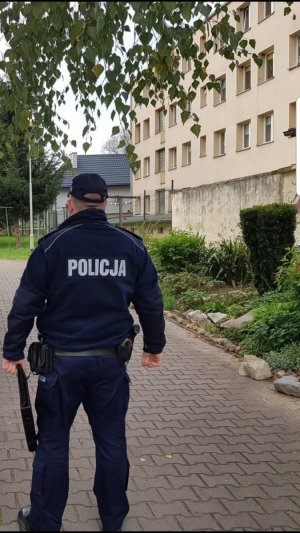 Policjant idzie  w kierunku budynku wielorodzinnego w tle widać budynek. Policjant trzyma  w ręku teczkę