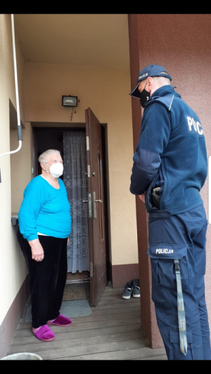 policjant stoi przy drzwiach wejściowych do domu, obok niego stoi kobieta. Osoby te mają maseczki na twarzach.