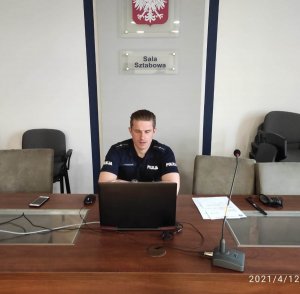 Policjant siedzi przy komputerze w sali sztabowej kłodzkiej komendy policji i uczestniczy w spotkaniu spotkanie senior online