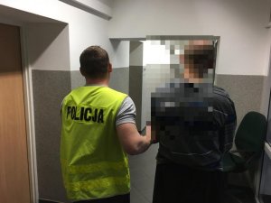 policjant prowadzi zatrzymanego mężczyznę, który ma kajdanki na rękach. Zdjęcie wykonane w pomieszczeniu służbowym