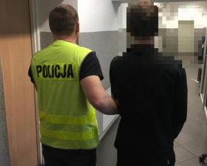 policjant prowadzi zatrzymanego mężczyznę, który ma kajdanki na rękach. Zdjęcie wykonane w pomieszczeniu służbowym