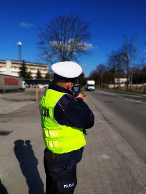 policjant z miernikiem prędkości dokonuje pomiaru prędkości samochodu jadącego jezdnią , na poboczu zaparkowany radiowóz policyjny