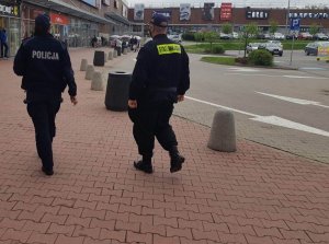 patrol policjant i strażnik miejski idą obok galerii handlowej w tel widać samochody oraz budynek handlowy galerii