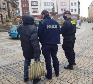 Patrol mundurowych policjant i strażnik miejski  rozmawiają z mężczyzną na terenie rynku , w tle widać fragment ratusza oraz plac przy nim